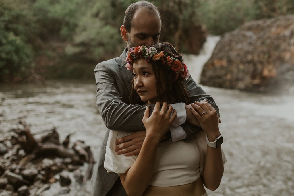Groom hugging Bride from behind by body of water