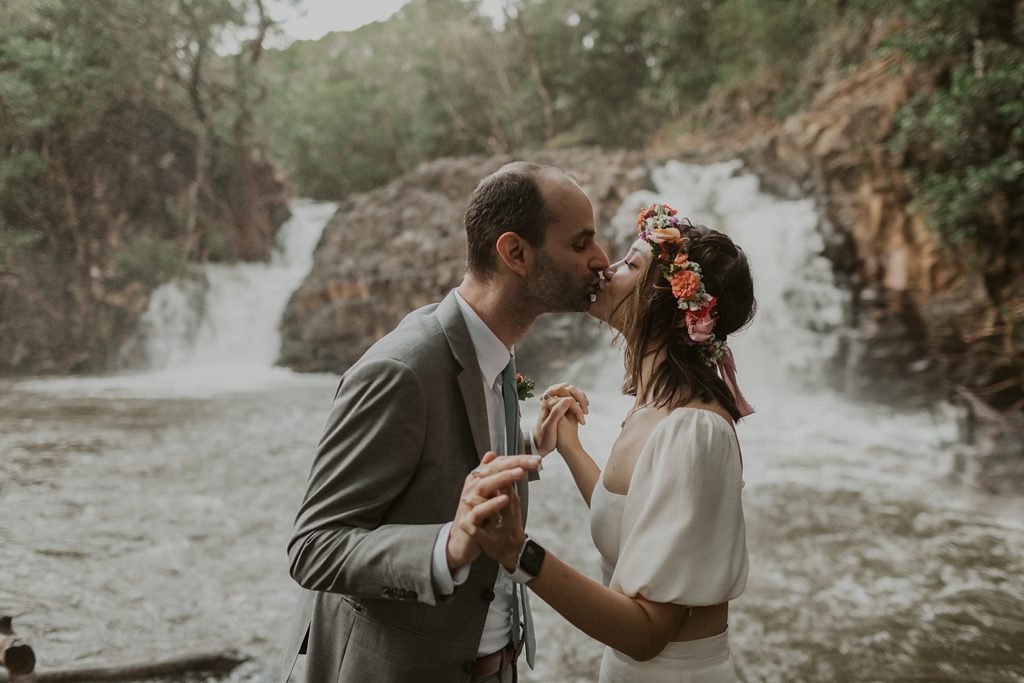 Bride and Groom holding both hands and kissing in front of waterfall