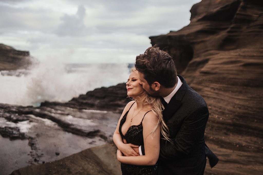 Groom holding and kissing Bride from behind by the ocean