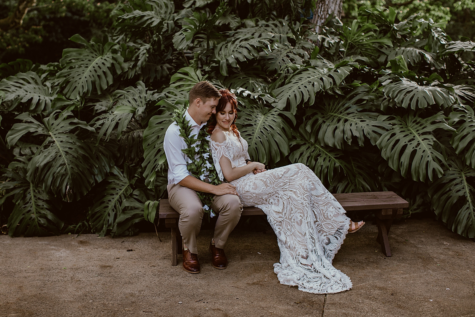 Groom sitting on bench with Bride leaning on him by green plants
