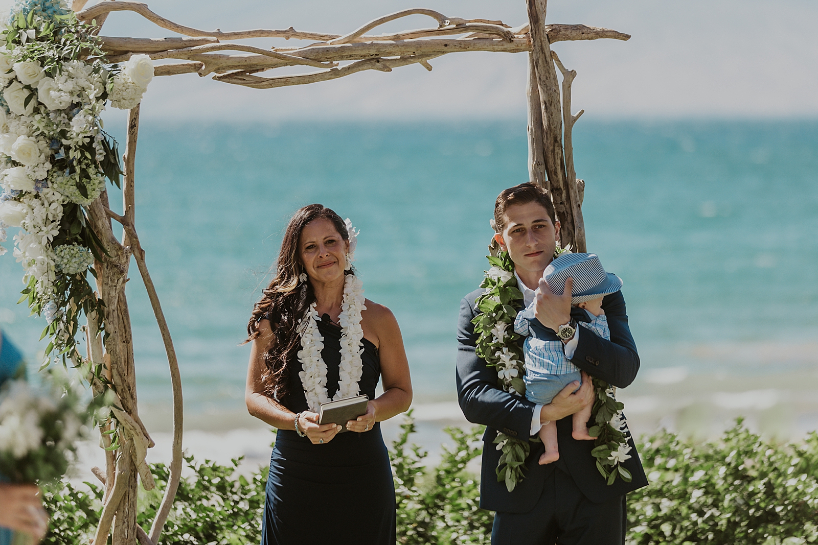 Groom holding baby by officiant at wooden branch arch by the water