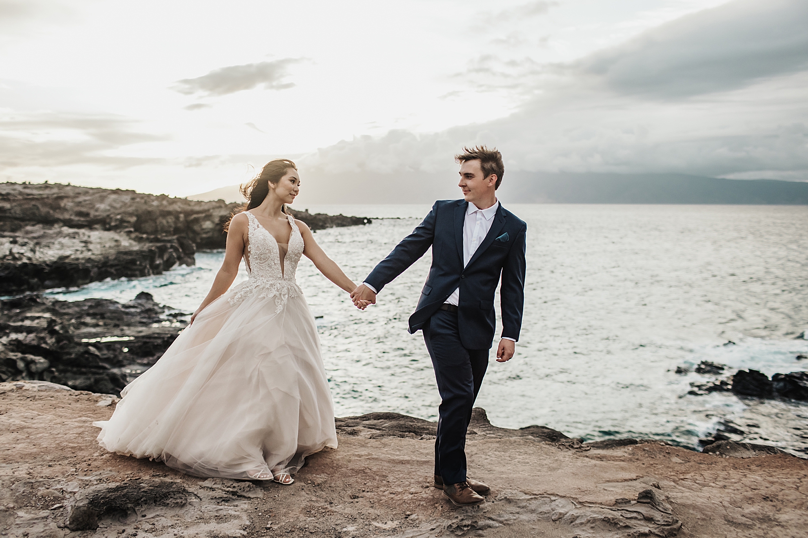 Groom holding Bride's hand leading by cliff side by the ocean