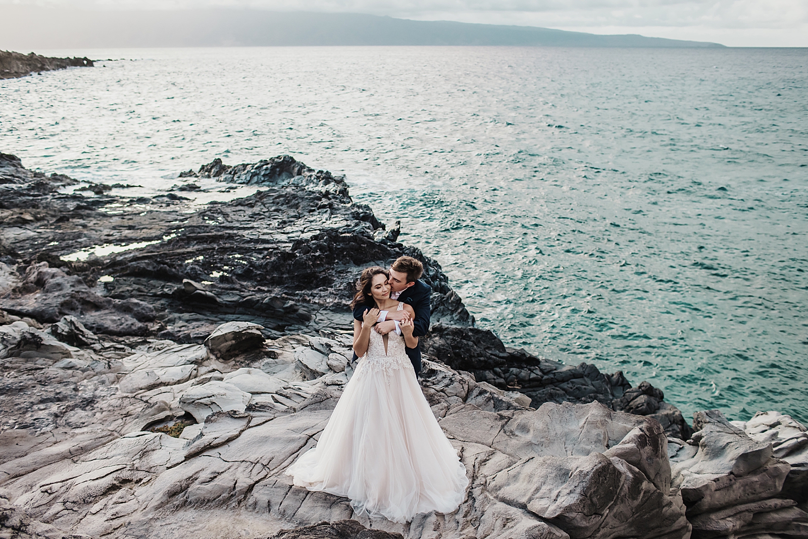 Groom holding Bride from behind and kissing her on rocky cliffside