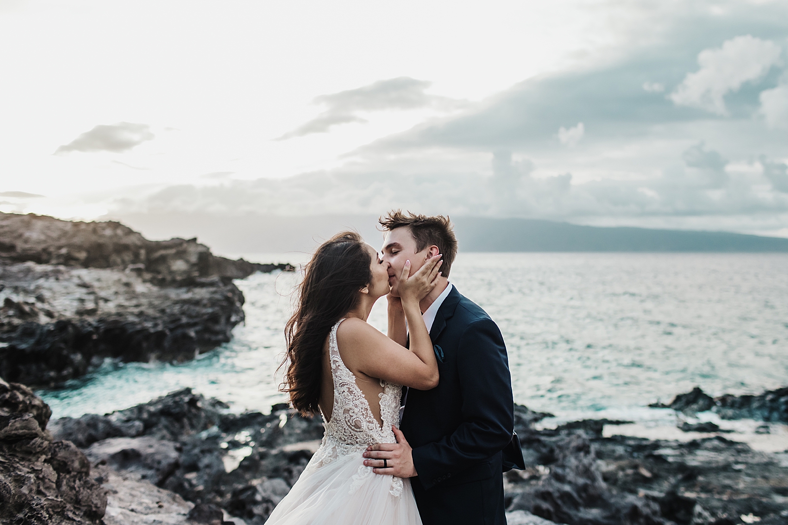 Bride holding Groom's face and kissing him on rocky ocean side