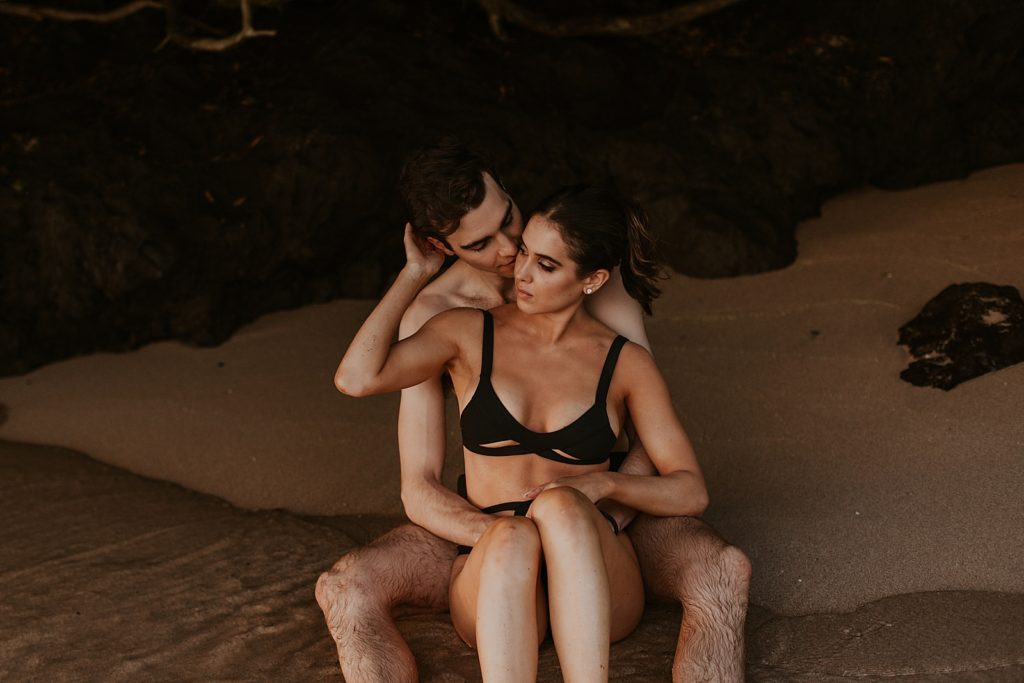 Woman sitting in man's lap and him kissing her while on the sand under land cover