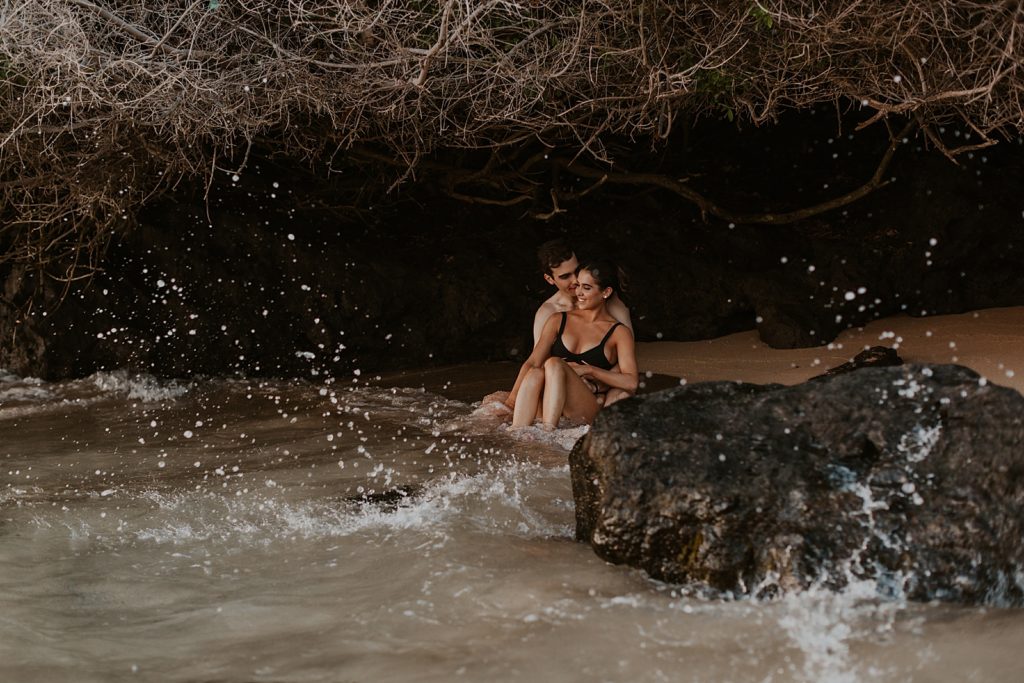 Couple sitting under land formation on the sand with water crashing in on the rocks