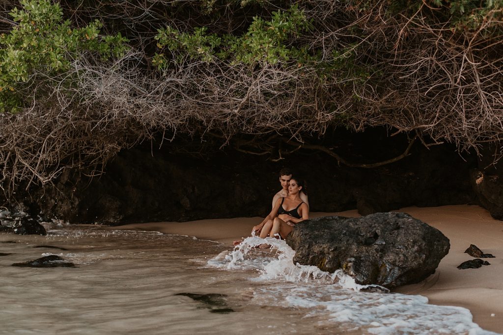 Couple sitting under land formation on the sand with water crashing in on the rocks