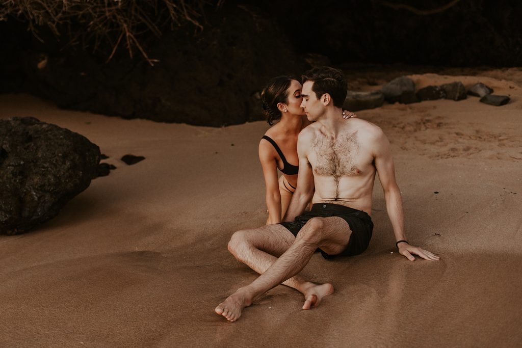 Woman kissing man's cheeck while sitting on the sand of the beach shoreline