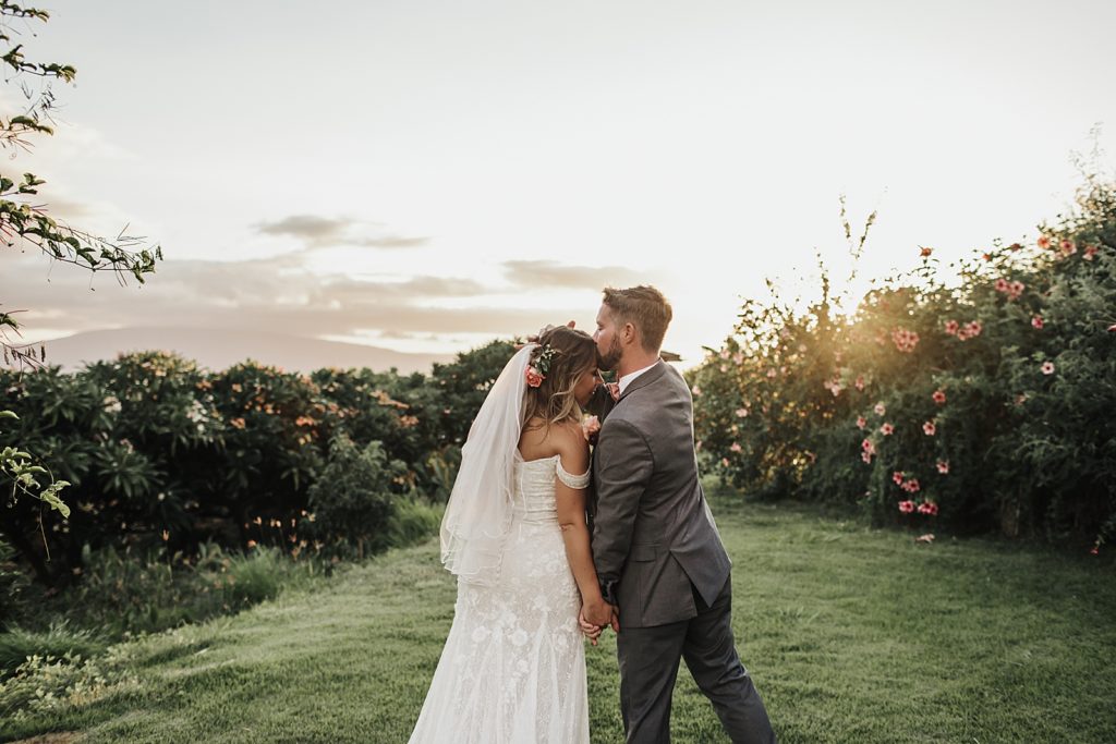 Groom kissing Bride on the forehead out on grassy field with pink flowers with the sun setting
