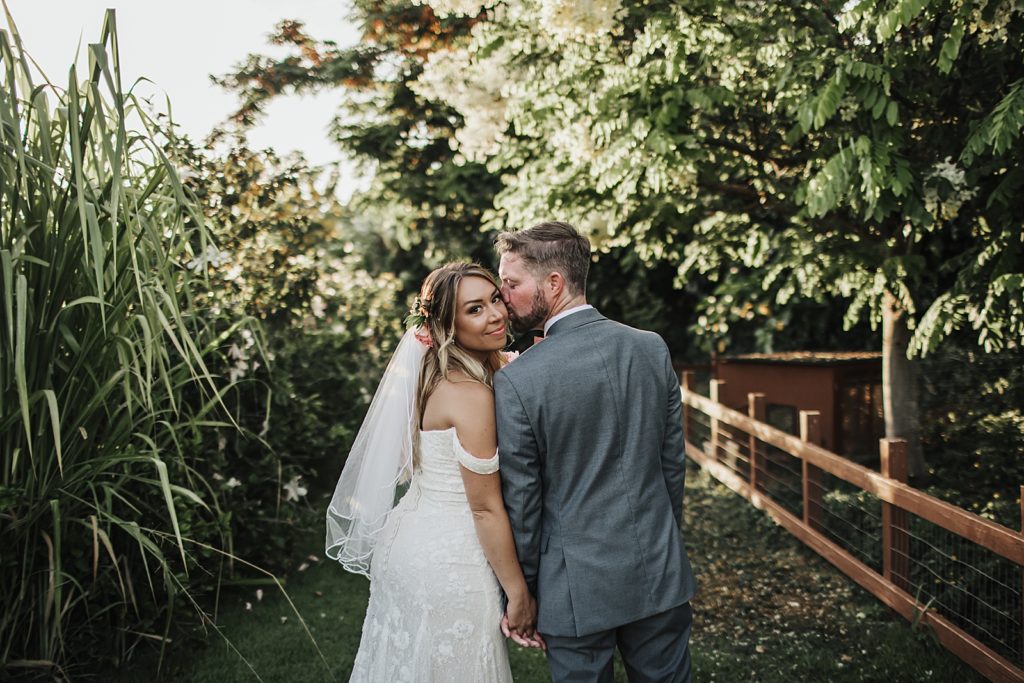 Groom kissing Bride on cheek as she looks behind surrounded by greenery