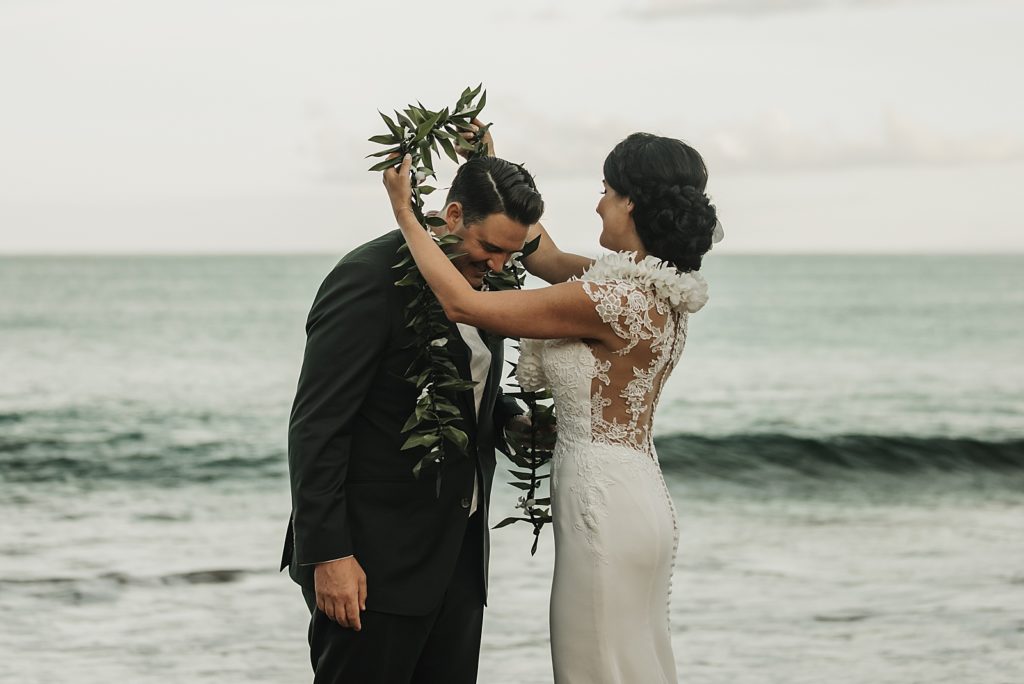 Bride putting greenery necklace around Groom's shoulders by the ocean