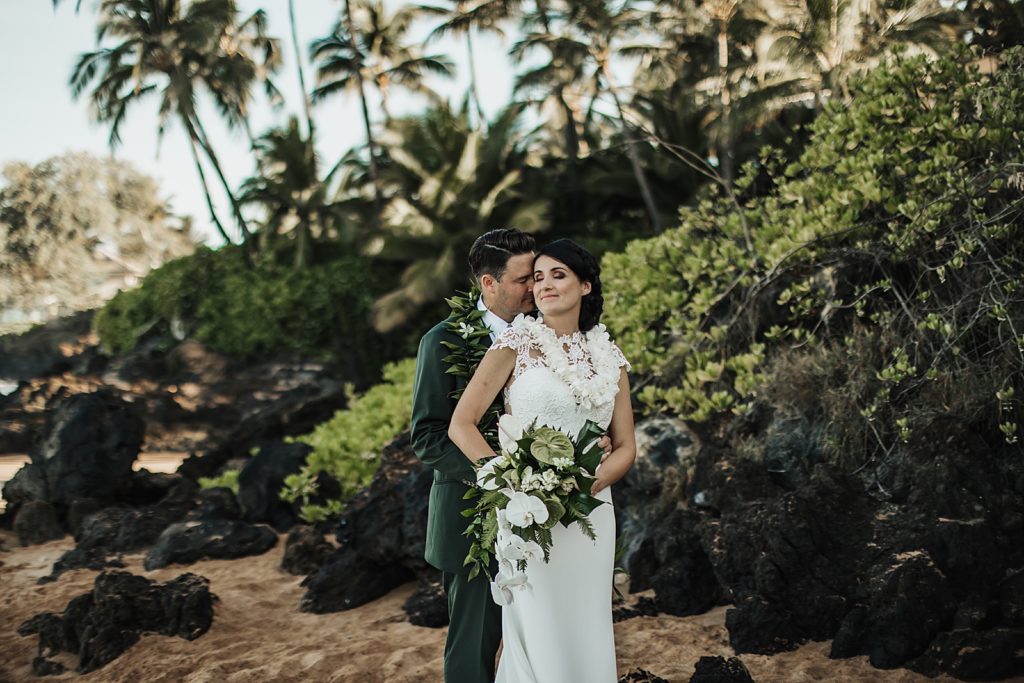Groom holding Bride and nuzzling by the greenery by the sand