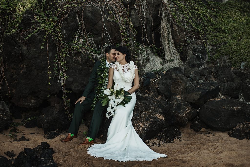 Groom resting on Bride's face while they sit on rocks by the beach