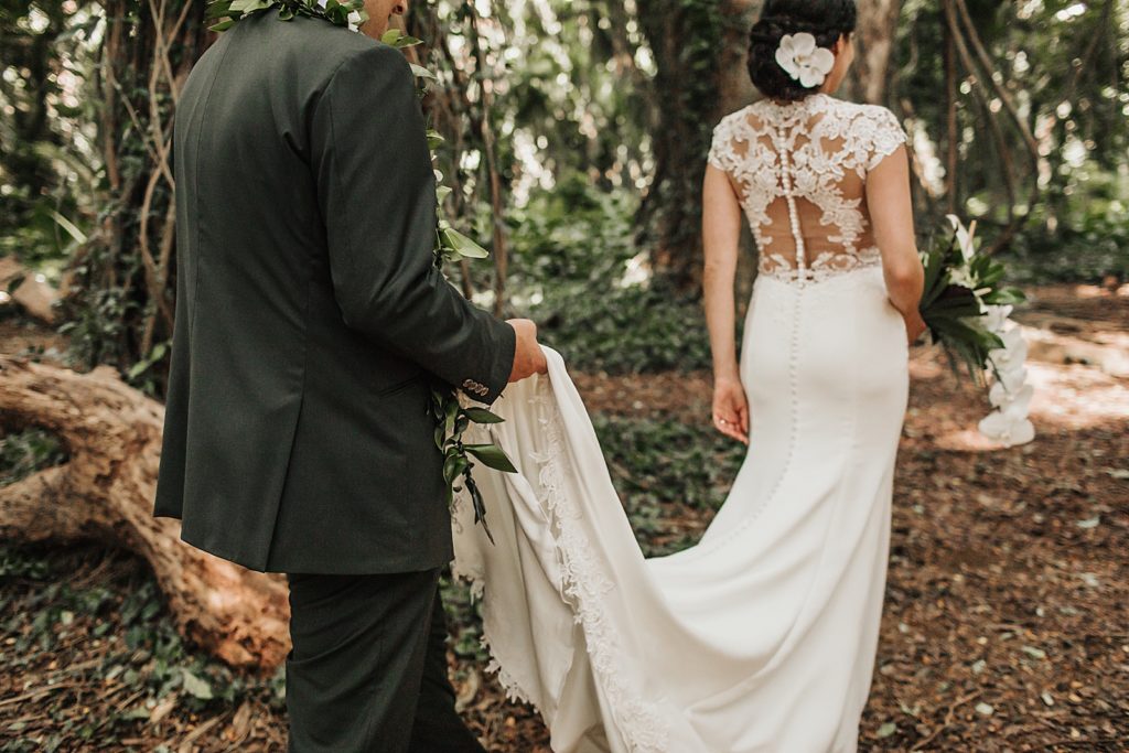 Closeup of Groom holding dress train assisting bride while walking in the forest