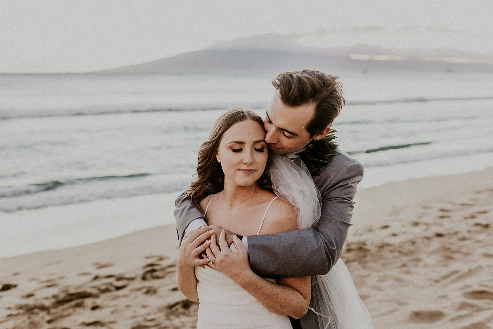 Groom holding Bride from behind on the beach by the ocean