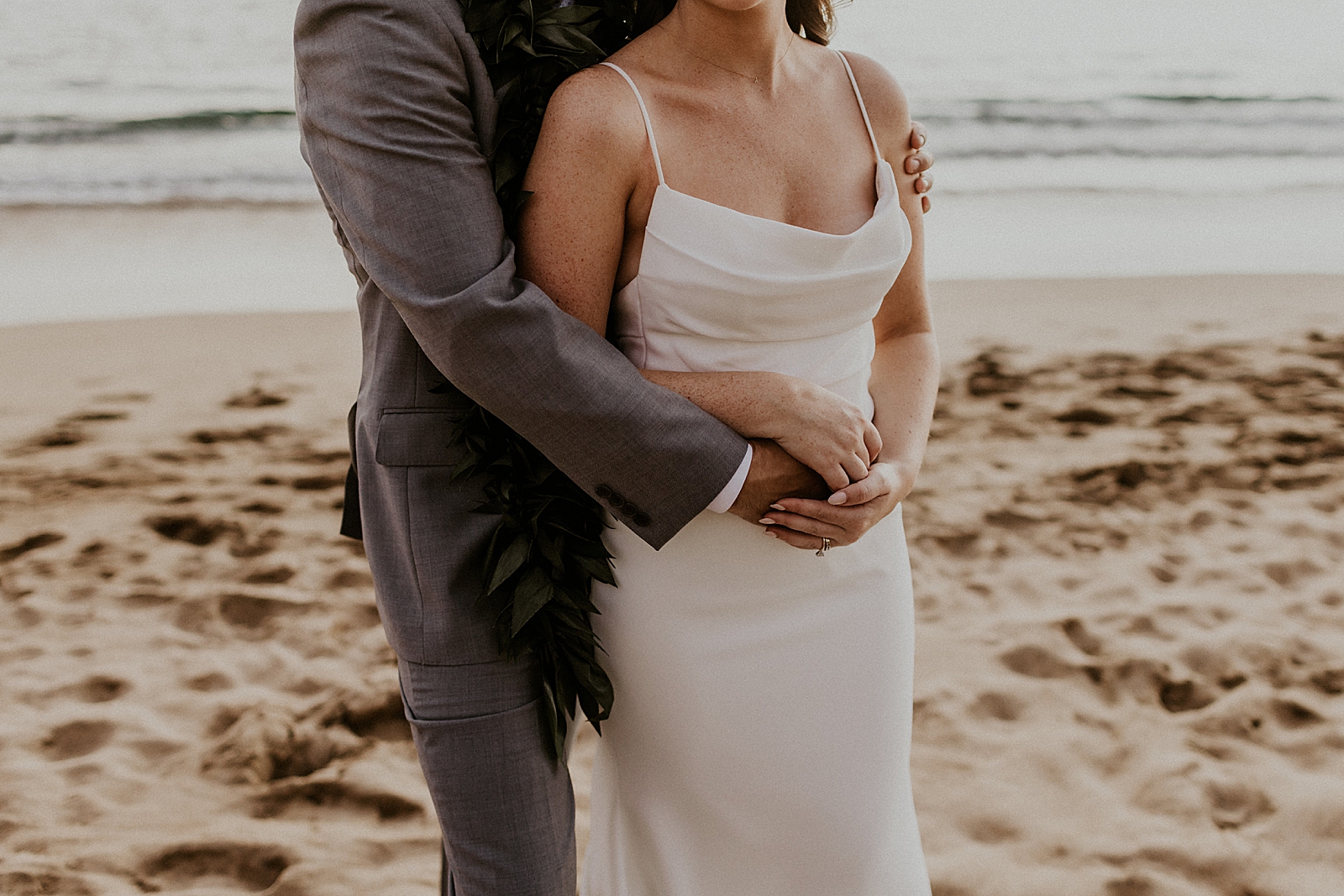 Mid shot of Groom hugging Bride from behind on the beach with the tides coming in