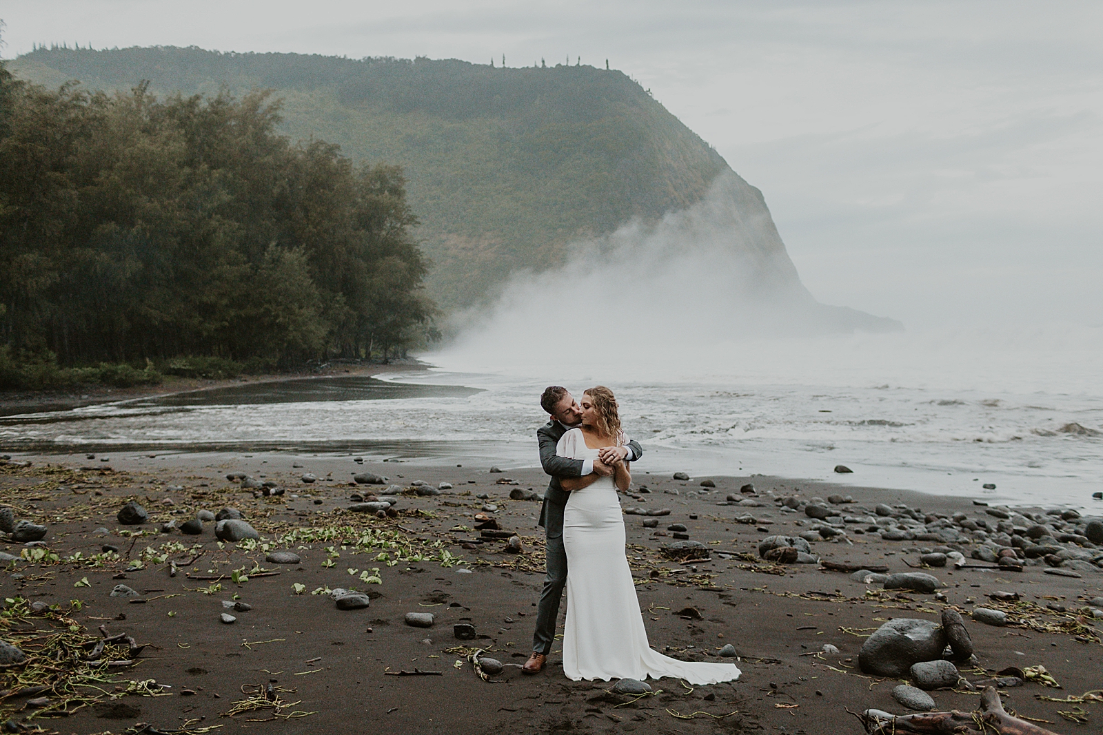 Groom holding Bride on the dark sand beach by the ocean with fog