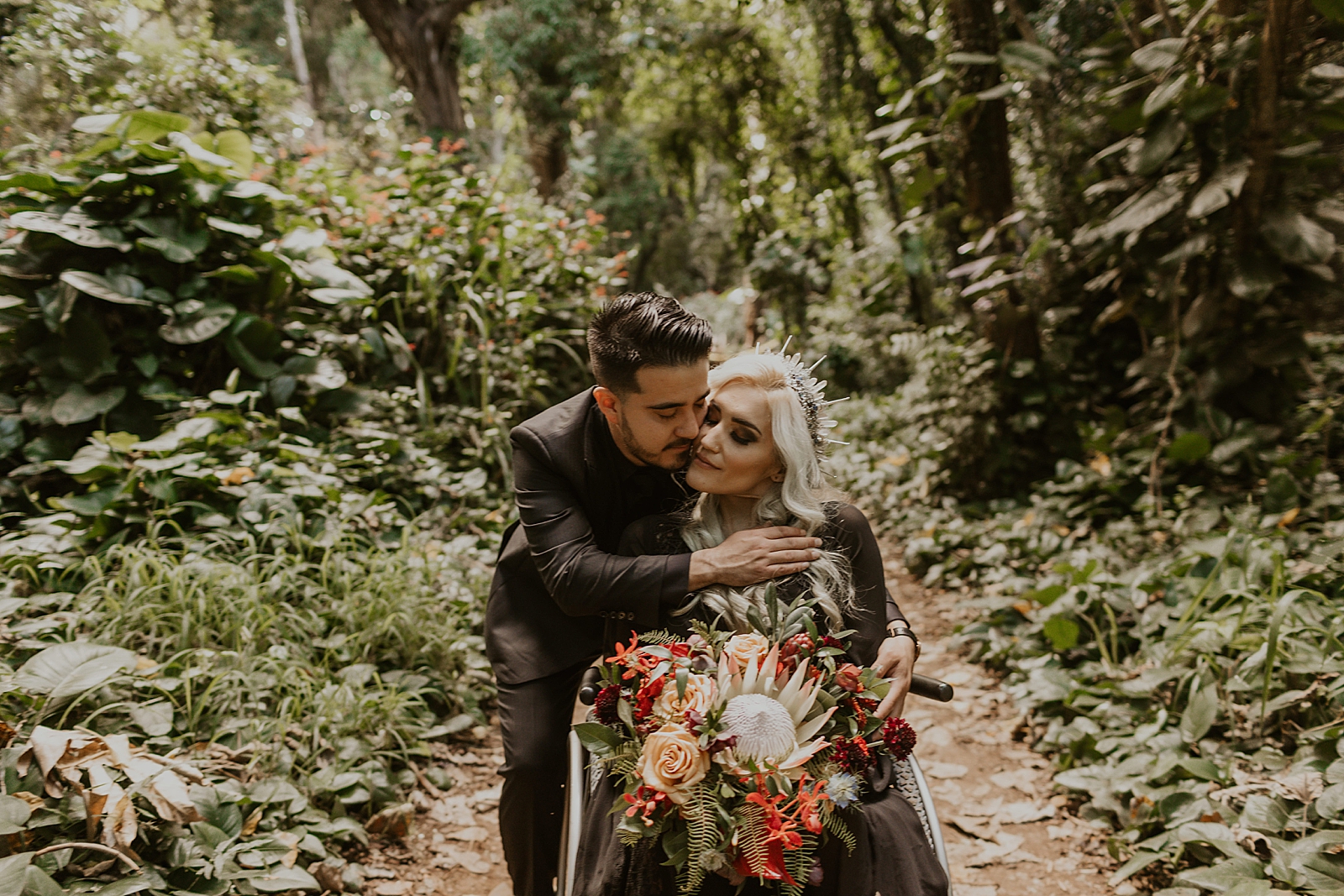 Groom leaning down and hugging wheel chaired Bride with bouquet in tropical forest 