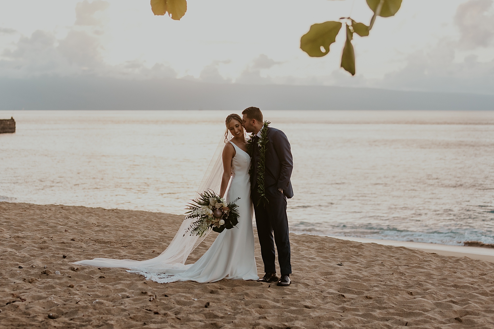 Groom kissing Bride on the cheek on the beach by the ocean