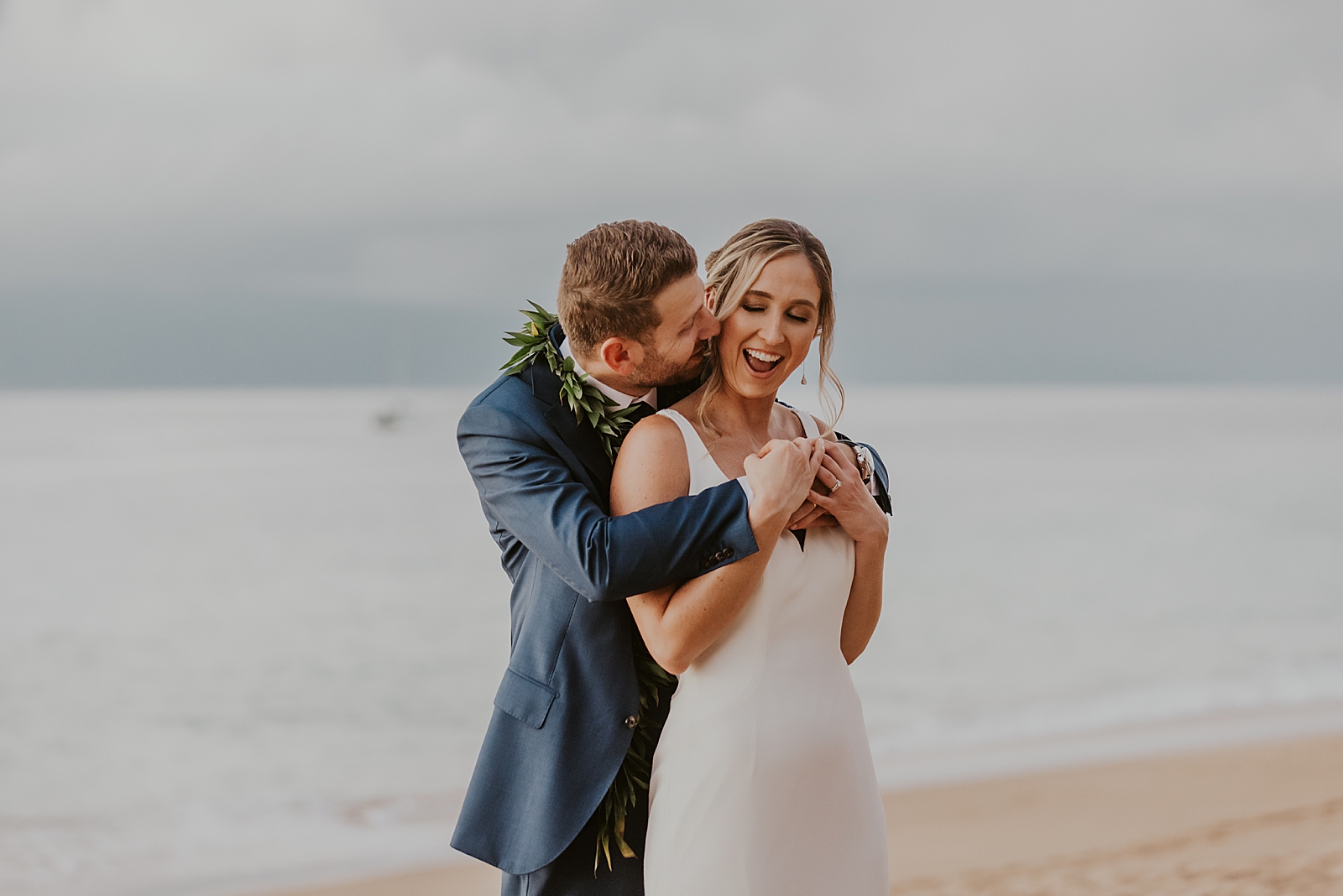 Groom holding Bride from behind and kissing her on the cheek on the beach