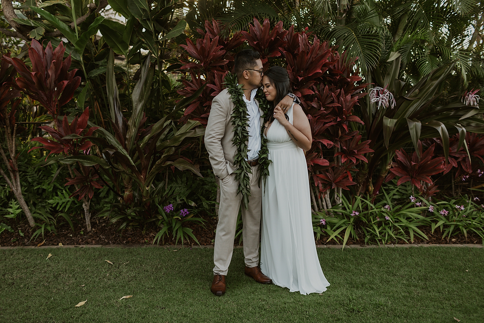 Groom kissing Bride on the forehead in front of greenery