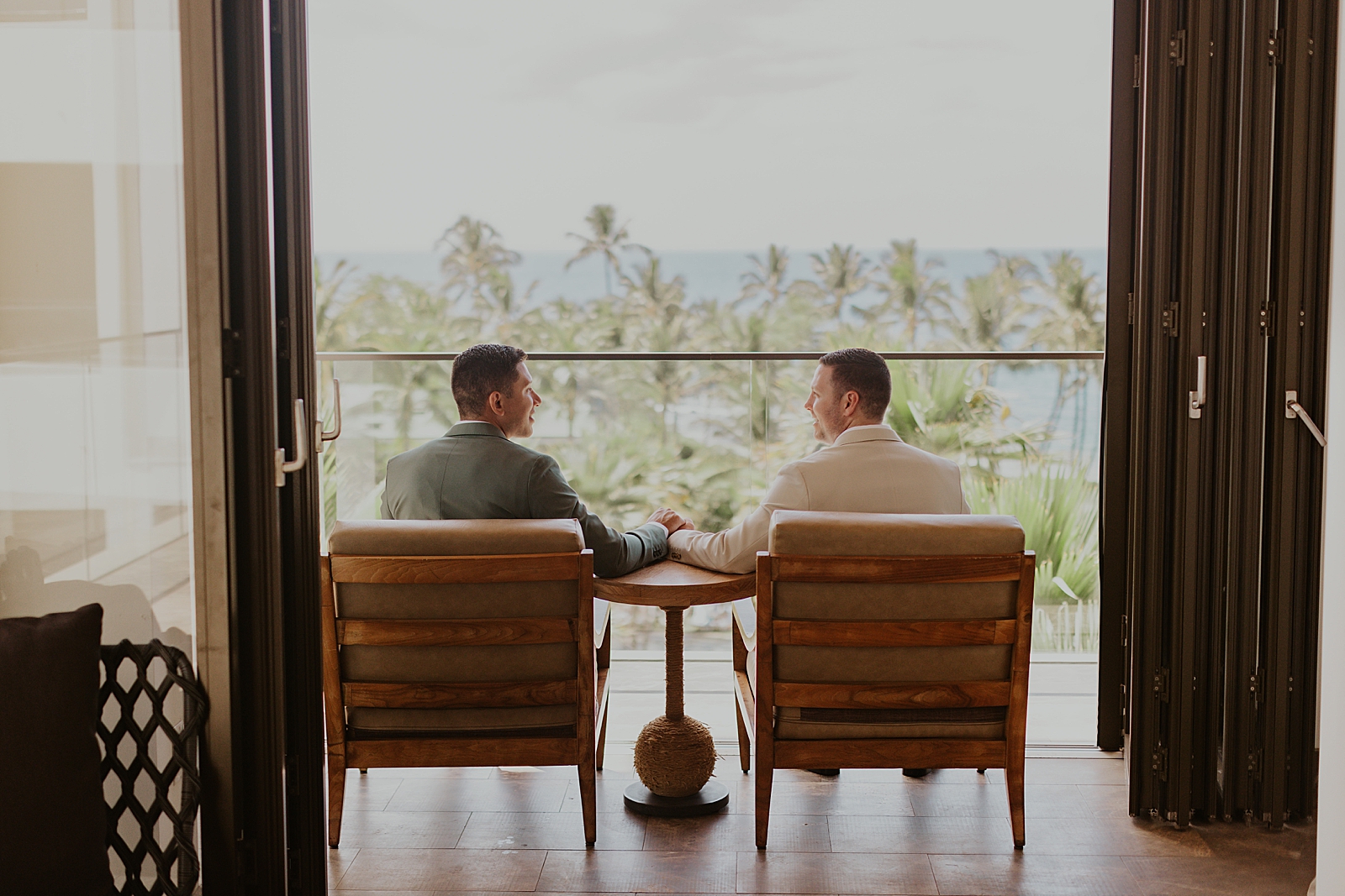 Grooms holding hands sitting on chairs looking out at the beach with palm trees