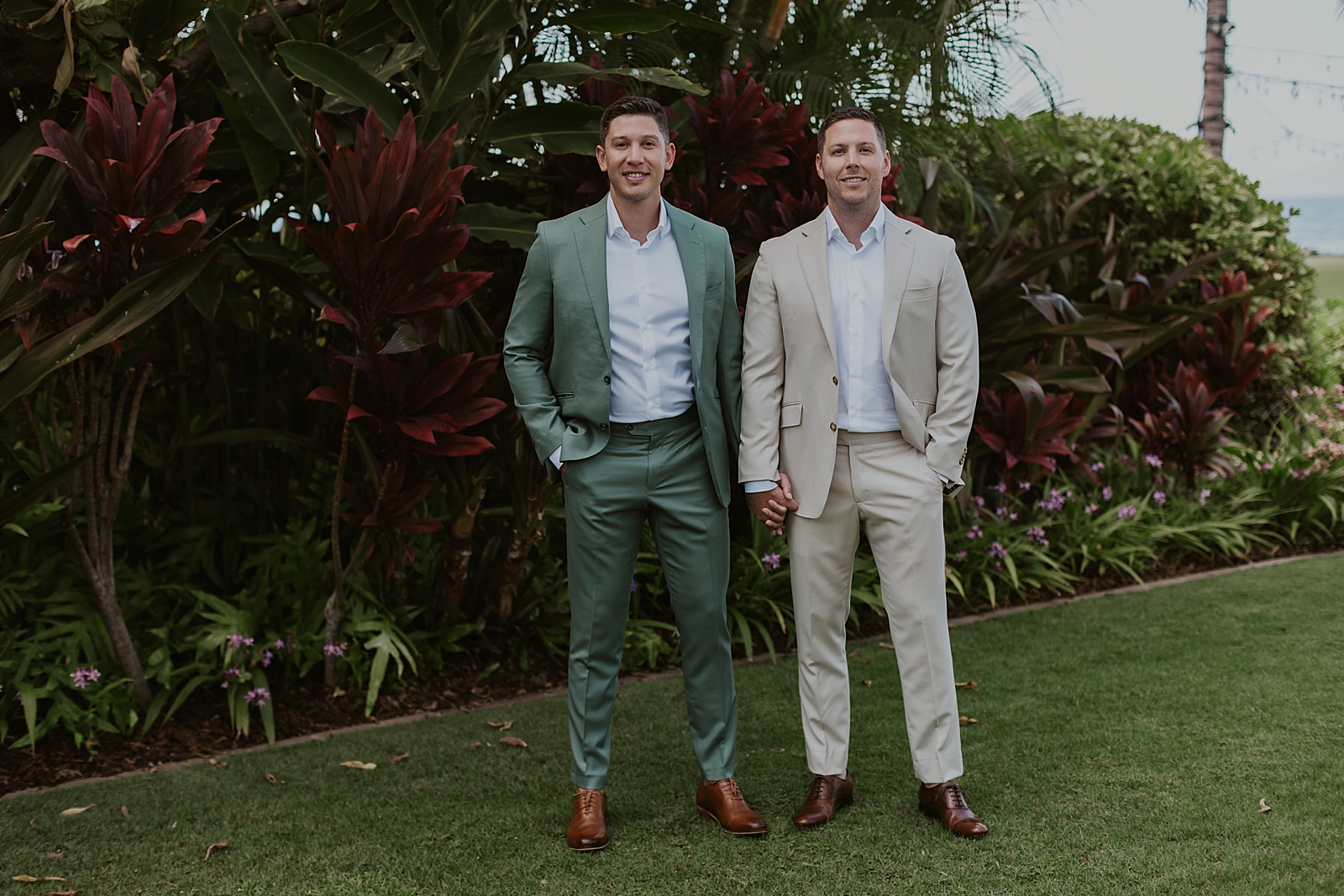 Grooms holding hands outside on green in front of tropical plants