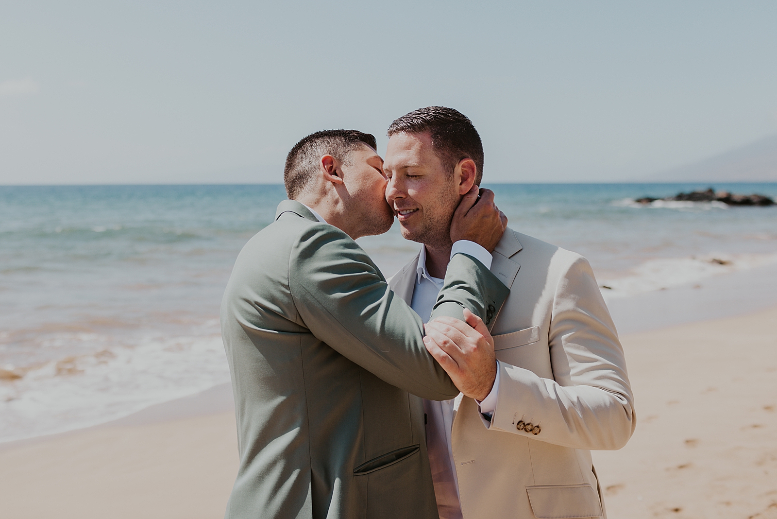 Groom kissing Groom on the cheek while standing out on the beach
