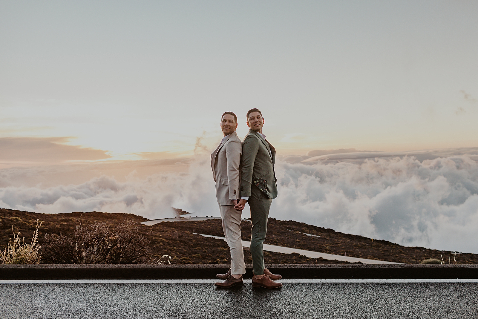Grooms back to back standing on mountain road with view of the top of the clouds