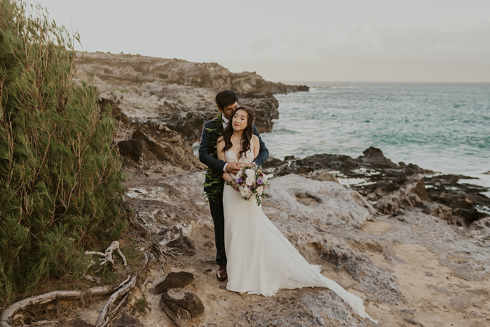 Groom hugging Bride from behind in front of the ocean