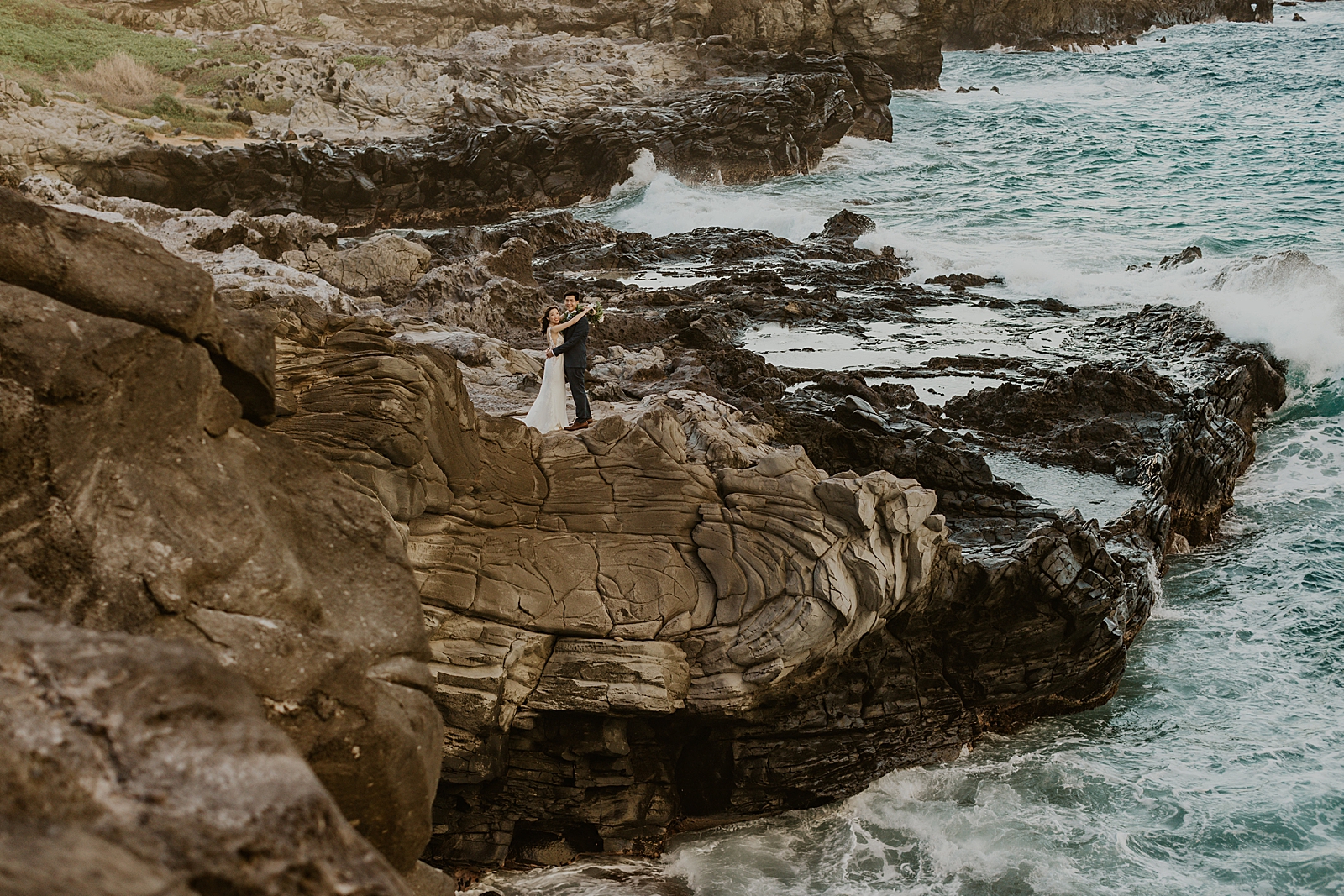 Wide shot of Bride and Groom holding each other by rocky cliffside of the ocean