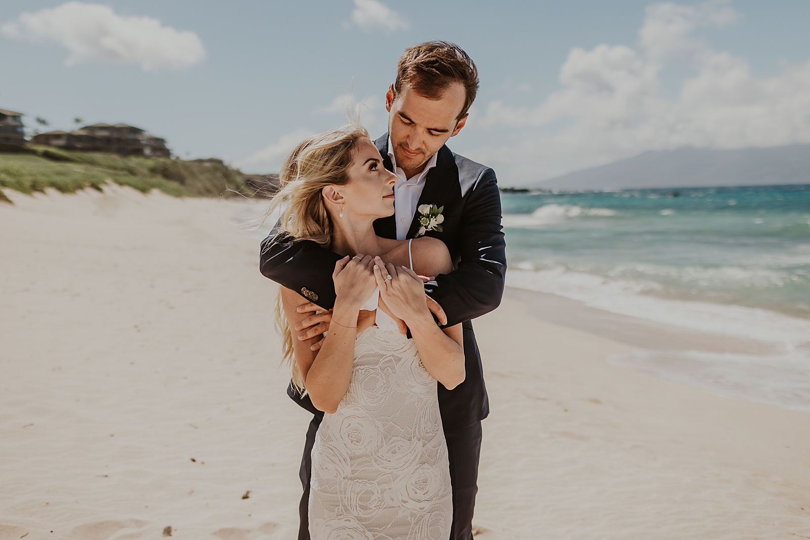 Groom holding Bride from behind on sunny beach with island in the distance