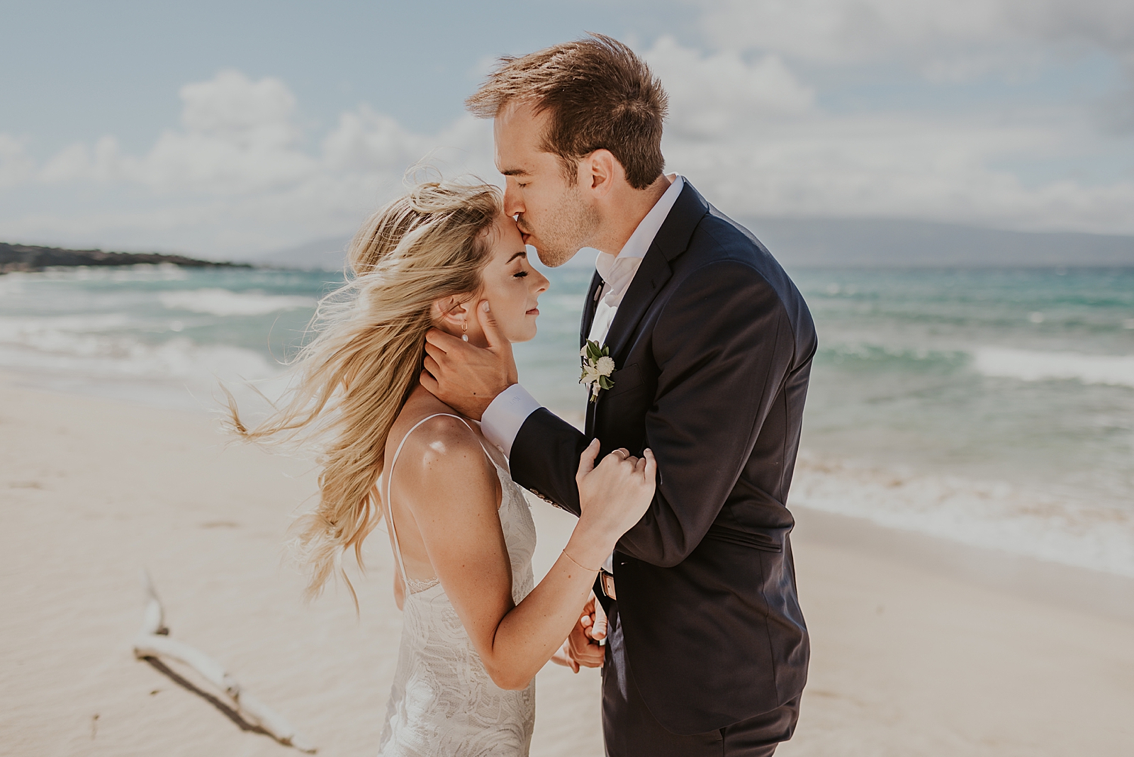 Groom holding Bride and kissing her on the forehead in front of the ocean